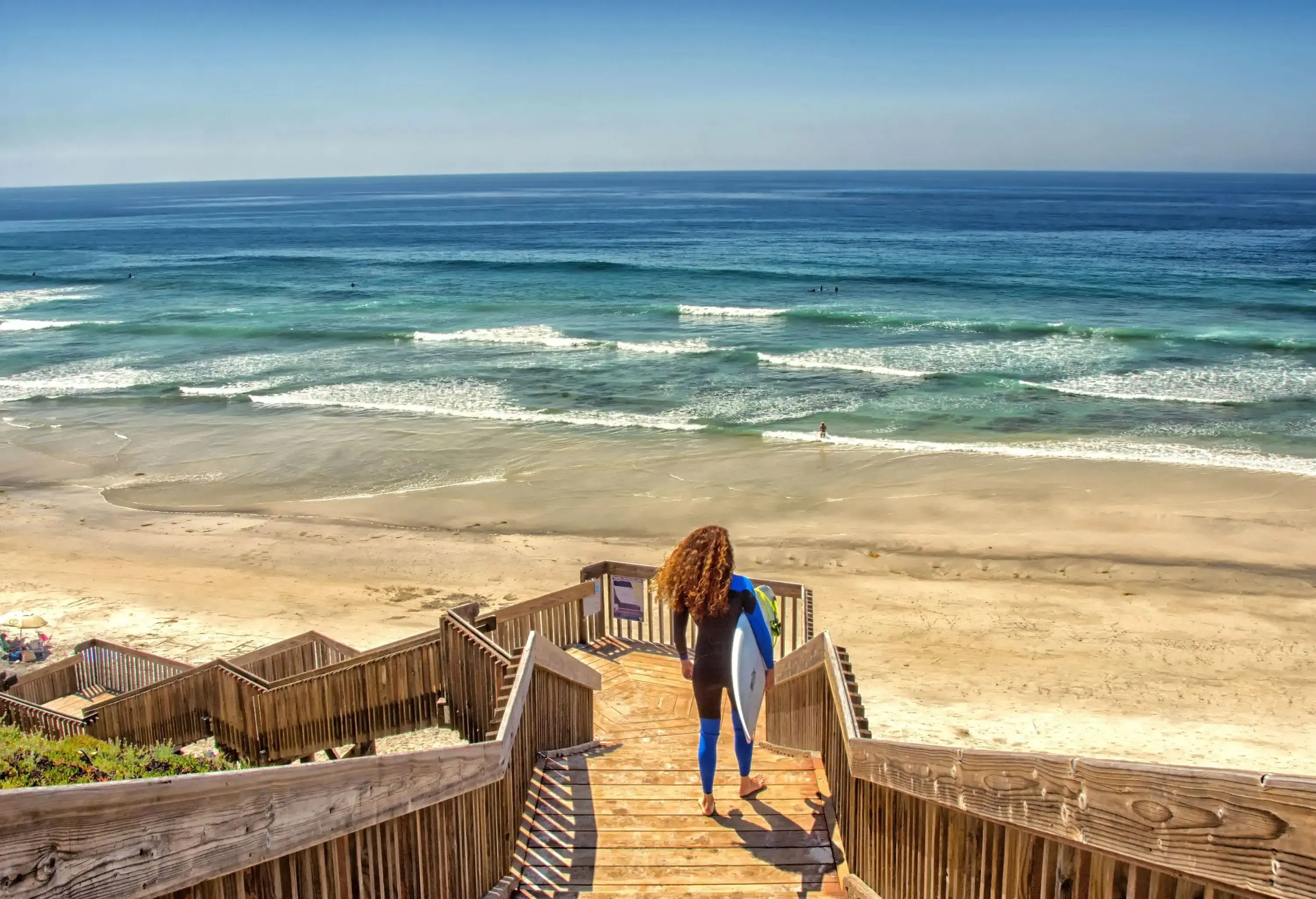 A surfer carrying a white surfboard walking down a long wooden staircase leading to the sea.