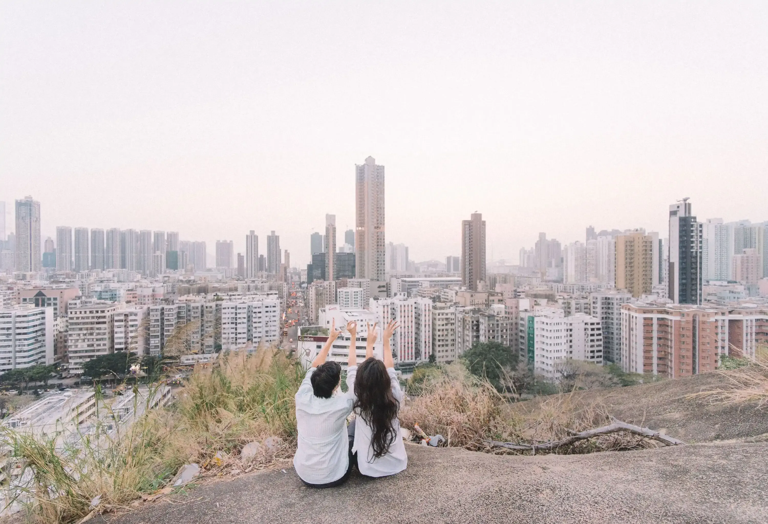 A sweet couple forms the love hand sign while sitting on the hill with the city skyline view against the white sky.