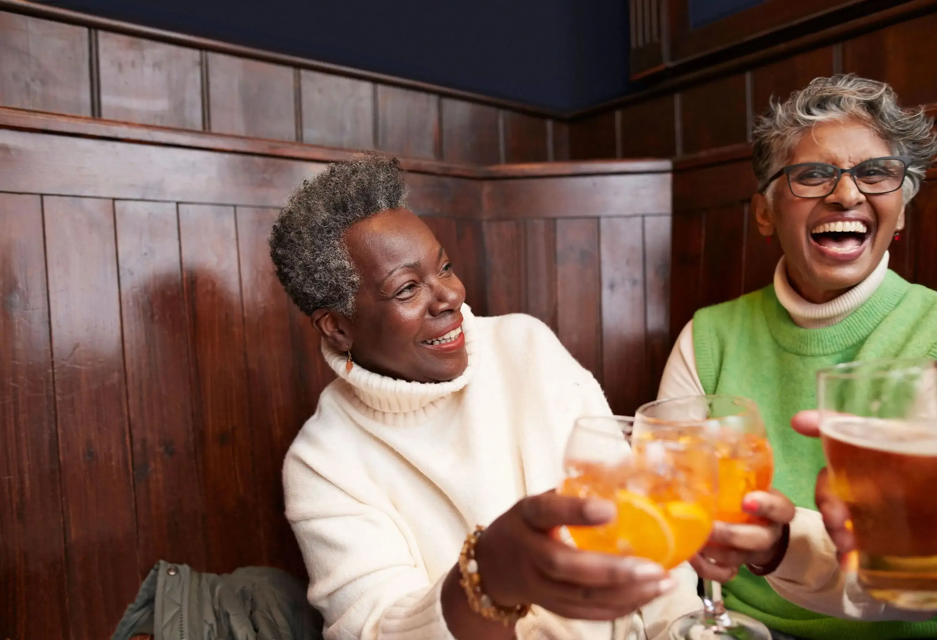 Two older women laughing and smiling together while holding drinks in a warm, wood-paneled room.