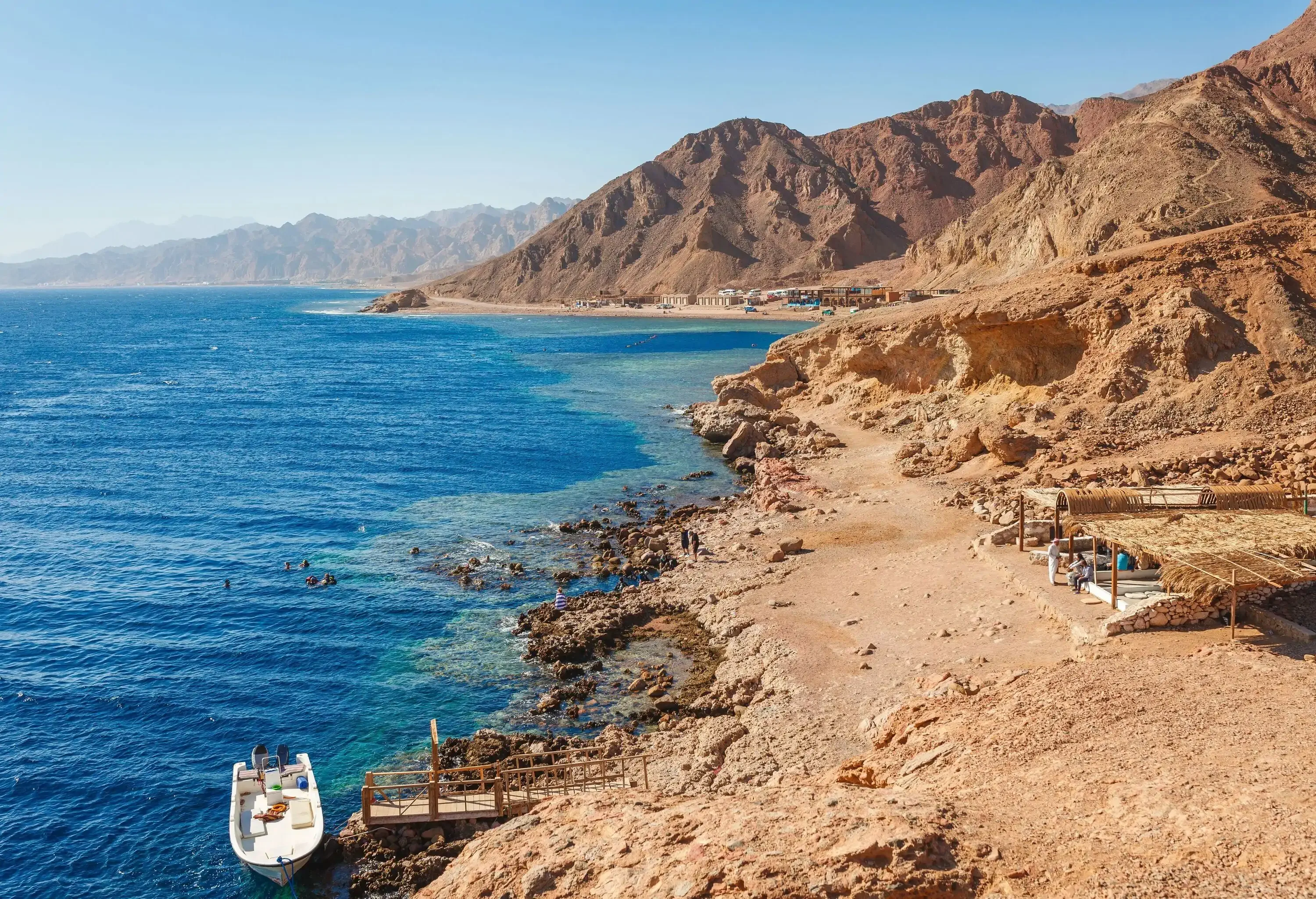 A white boat anchored beside a pier of a rugged beach with straw cottages along an eroded landscape.