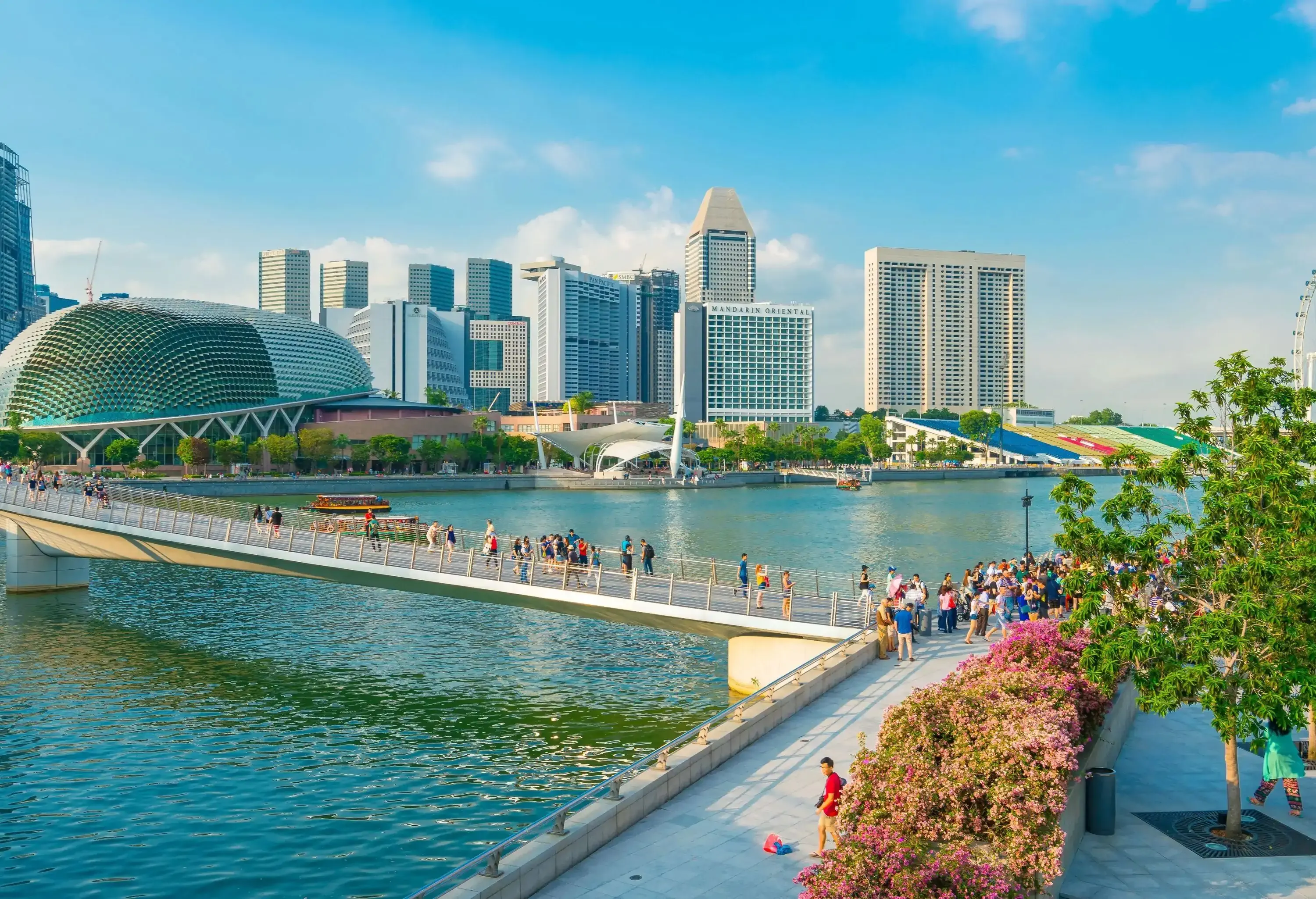 A city skyline on a bright day featuring a prominent pedestrian bridge over water in the foreground, leading to a cluster of modern buildings in the background.