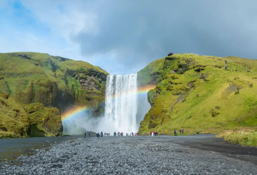 A mesmerising waterfall lined with a beautiful rainbow drops from a high cliff forming a lot of drizzle towards the visiting tourists in the foreground.