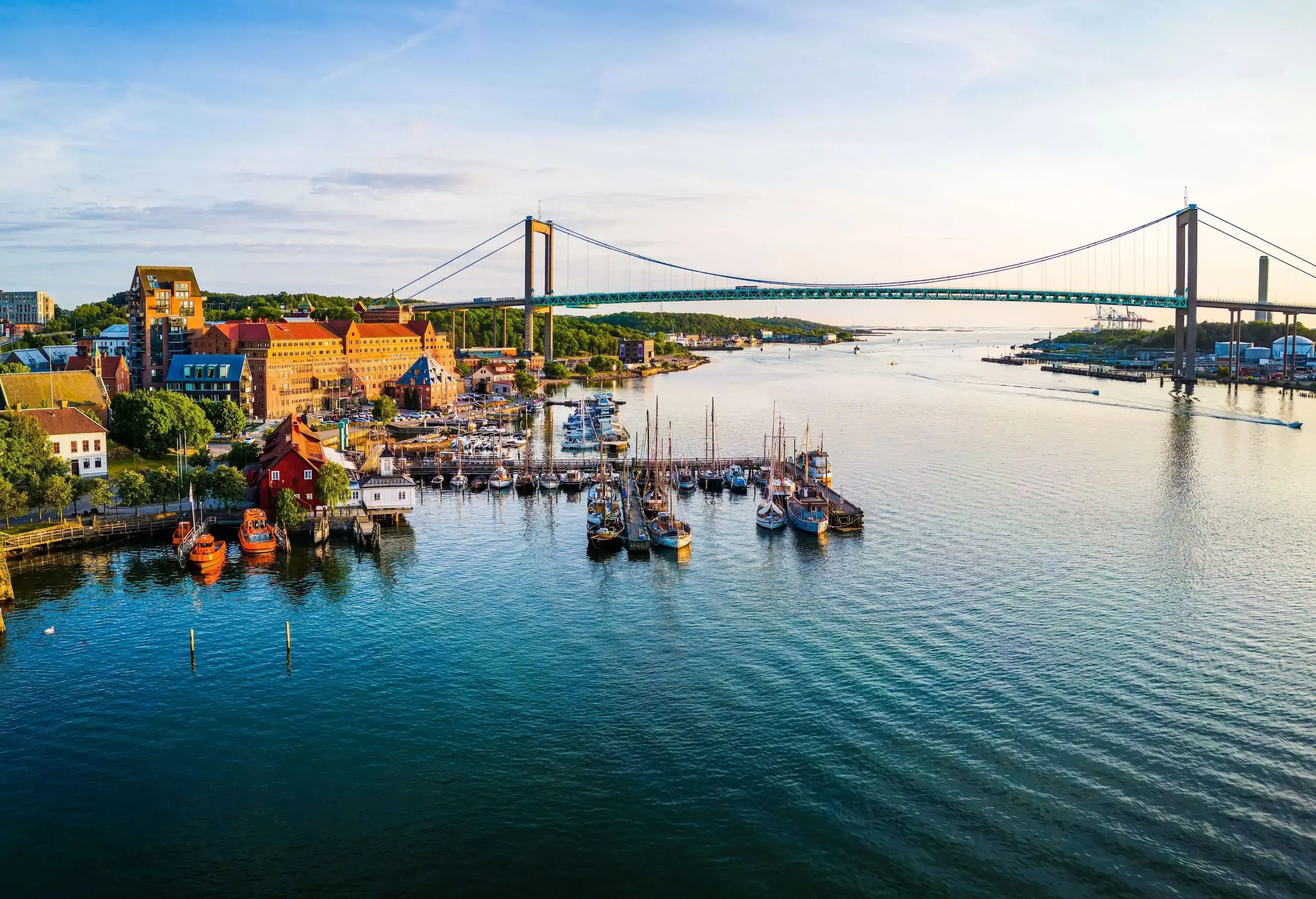 A grand suspension bridge spanning a calm body of water, with numerous boats docked in the foreground and diverse buildings lining the waterfront.