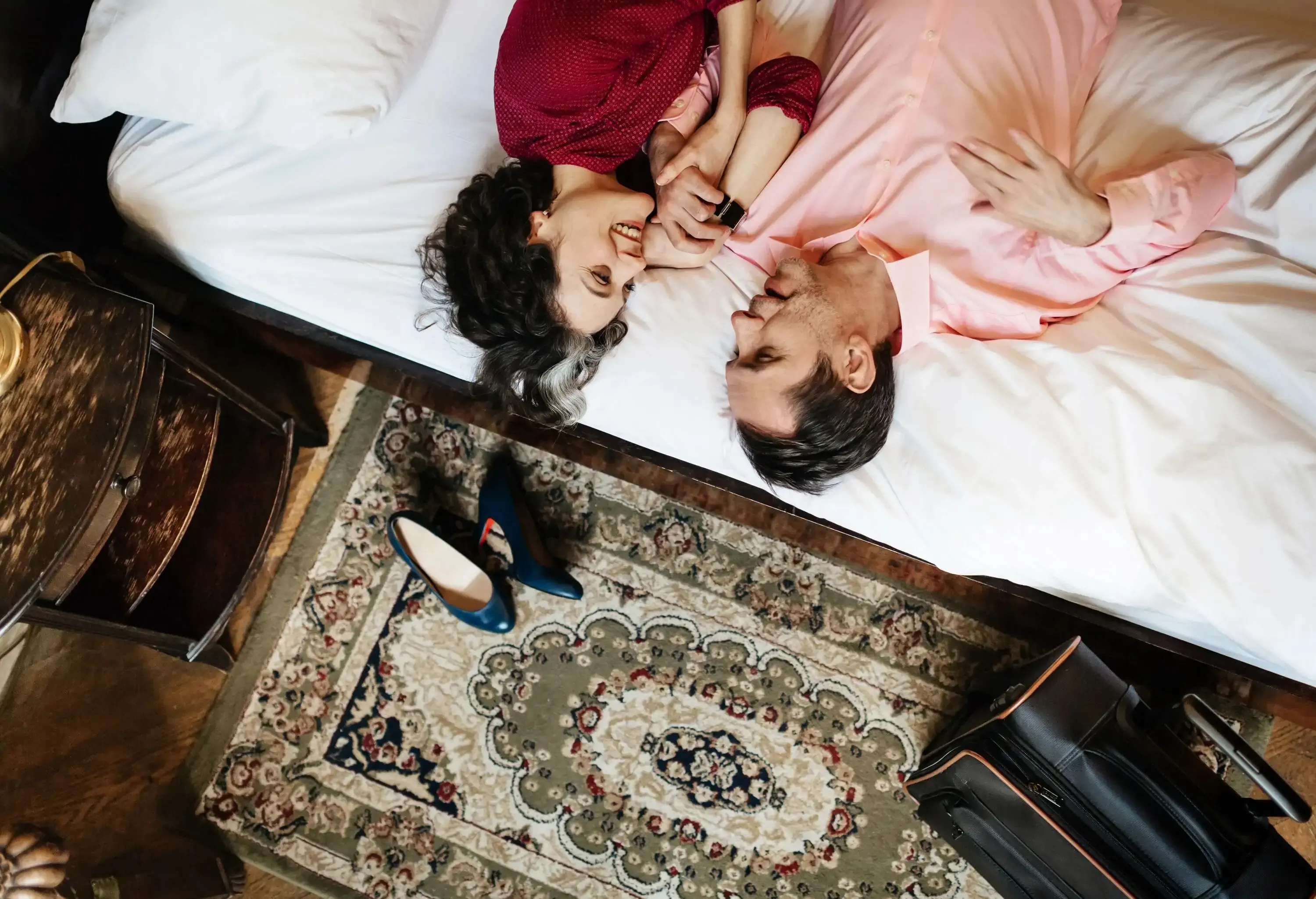 Overhead view of a smiling couple lying on a white bed, looking at each other, with a suitcase and a pair of blue high heels on a patterned rug below.