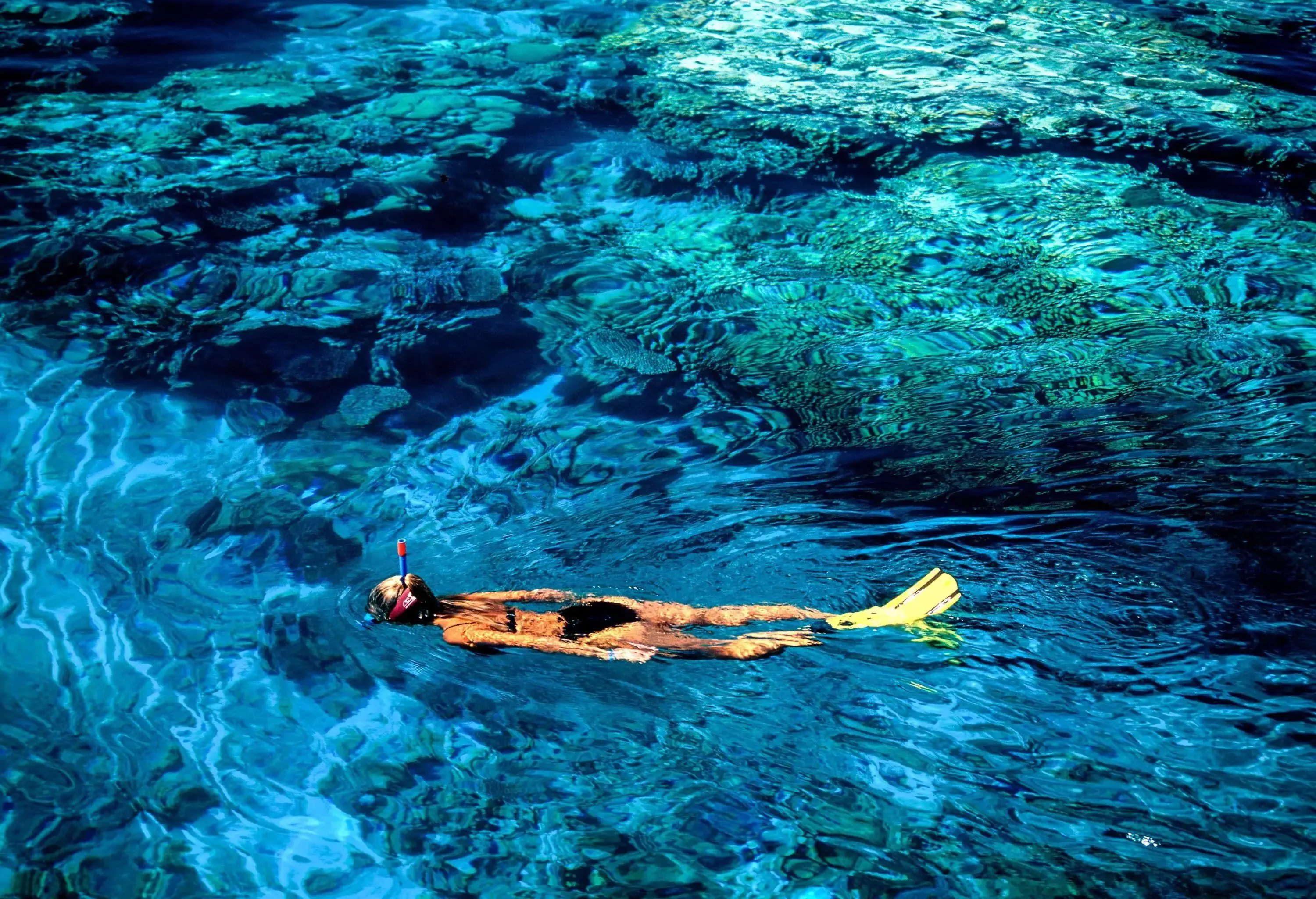 A woman in a black bathing suit snorkels across a crystal-clear water with coral reefs visible underwater.