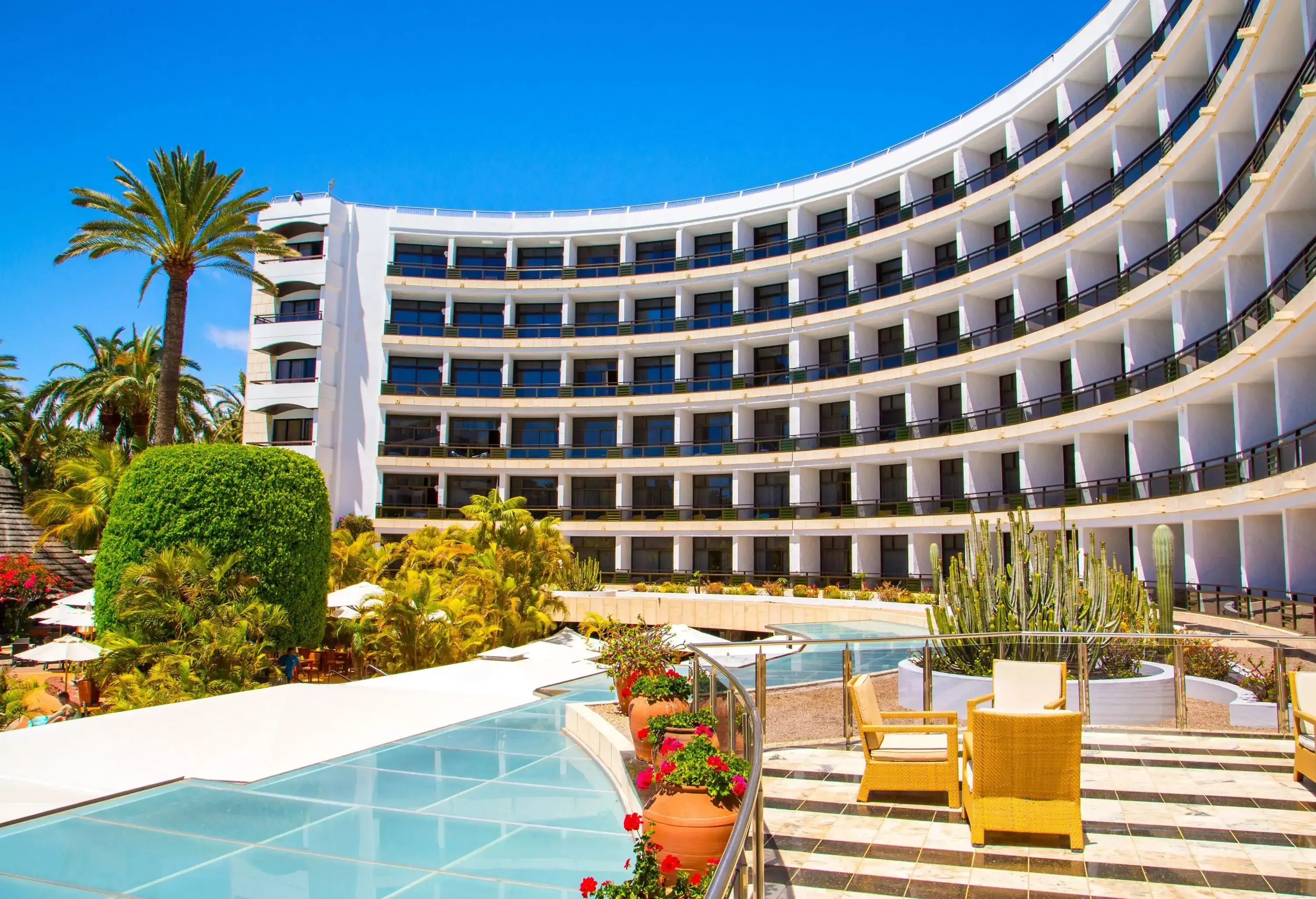 A crescent-shaped hotel building overlooking a swimming pool and a patio with chairs.
