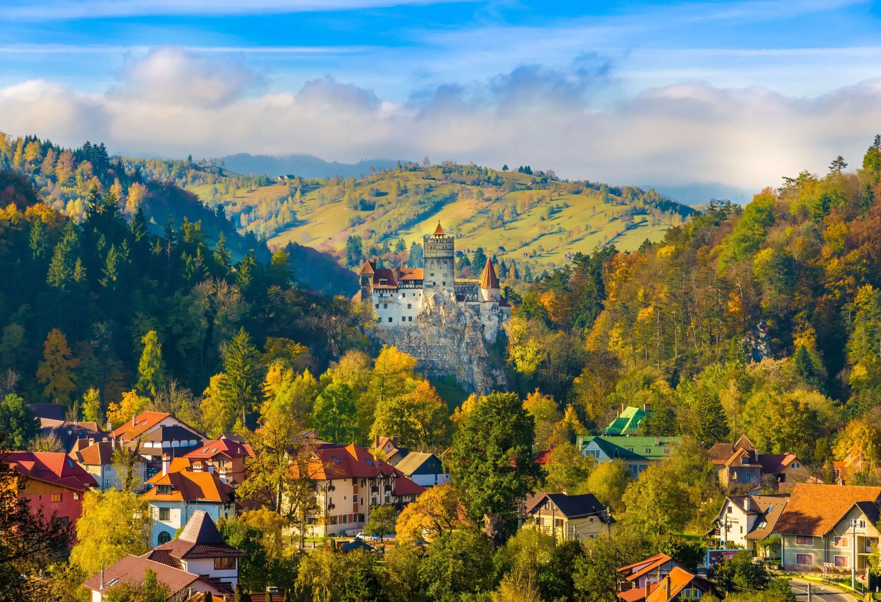 The medieval stone fortress Bran Castle, with a square-shaped turret, is perched on a rocky hilltop overlooking the cityscape surrounded by vibrant fall foliage.