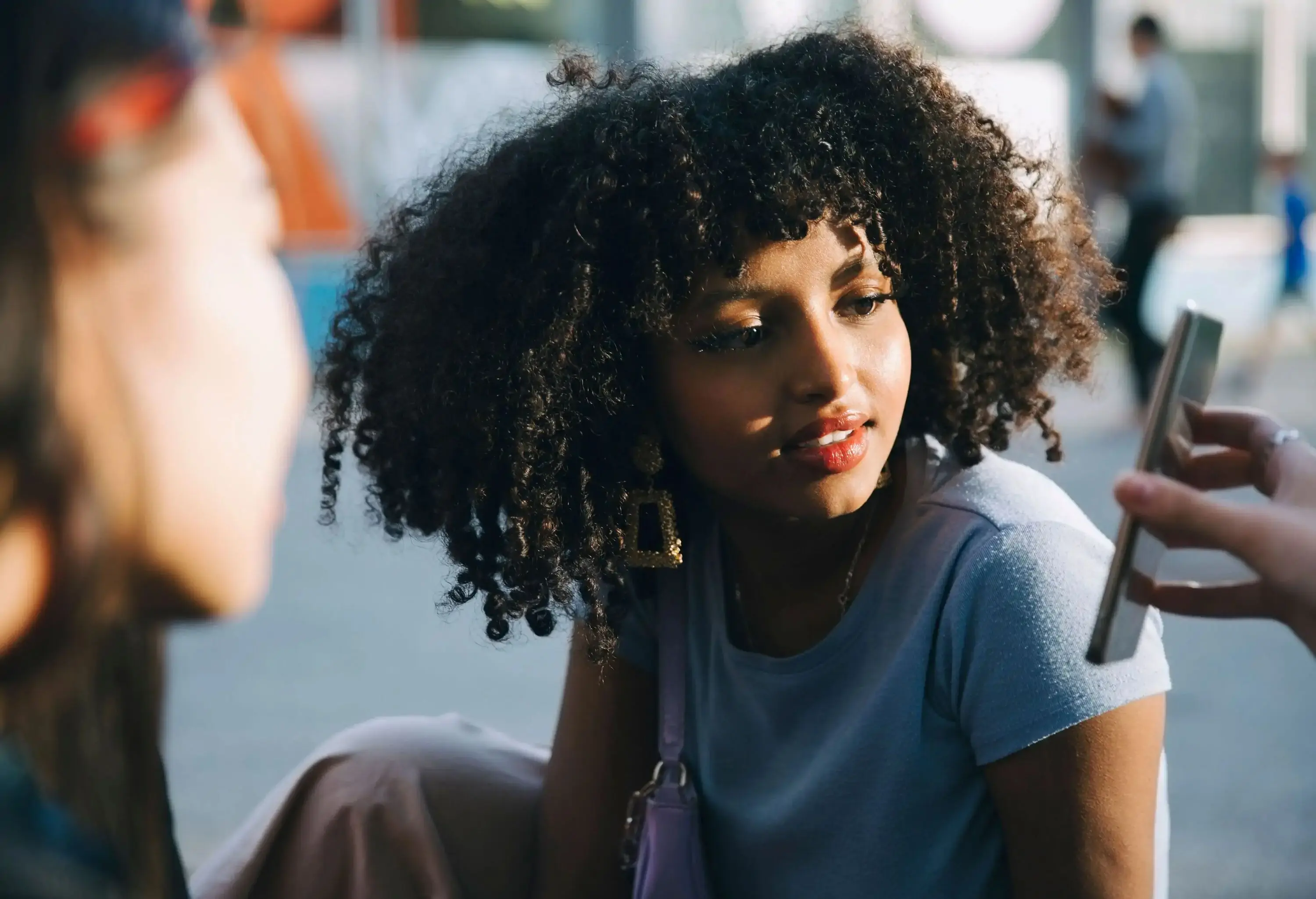 A young woman with curly hair staring at a smartphone that another person is showing her.