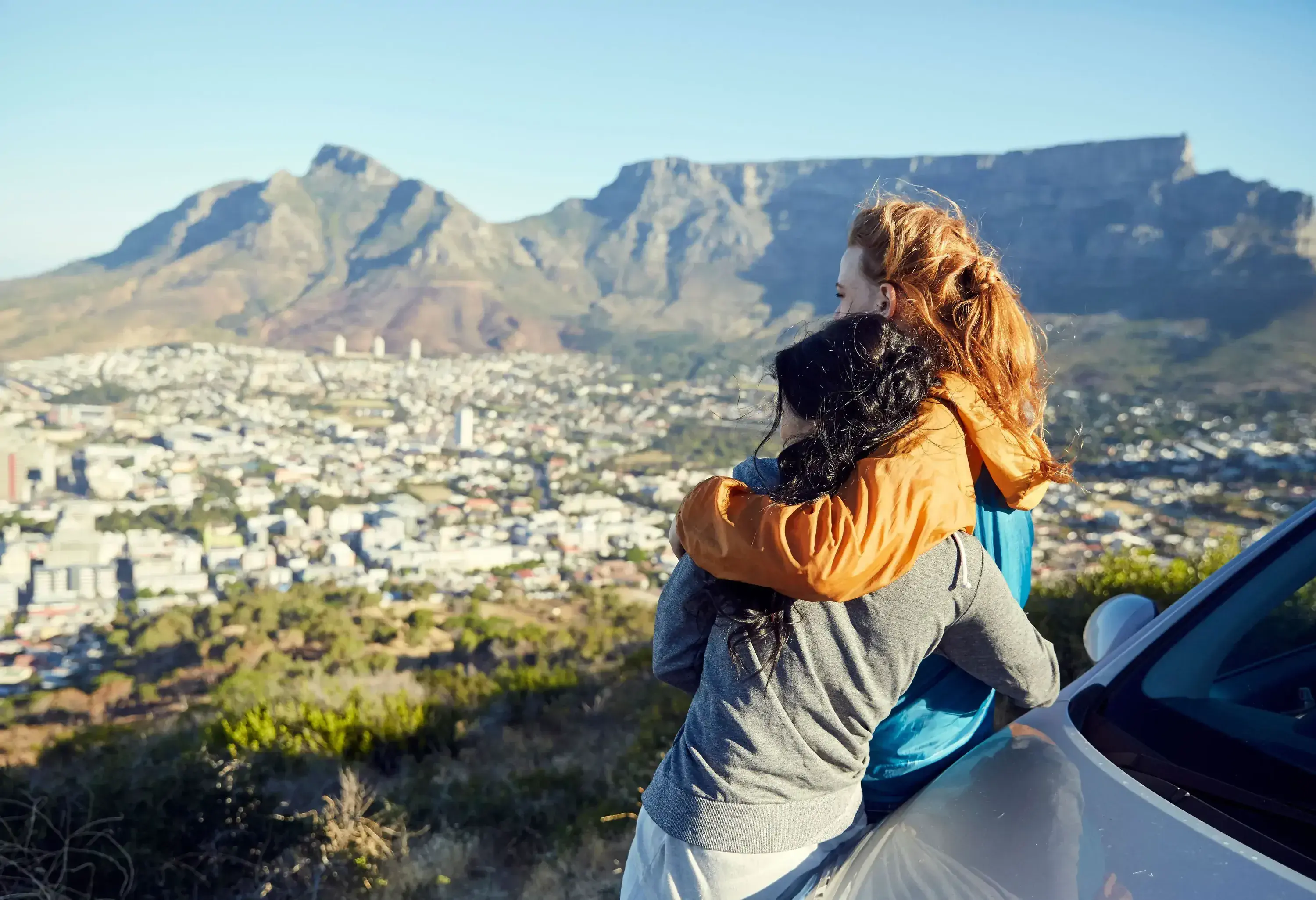 A female couple shares a hug beside a car as they look at a vast cityscape.