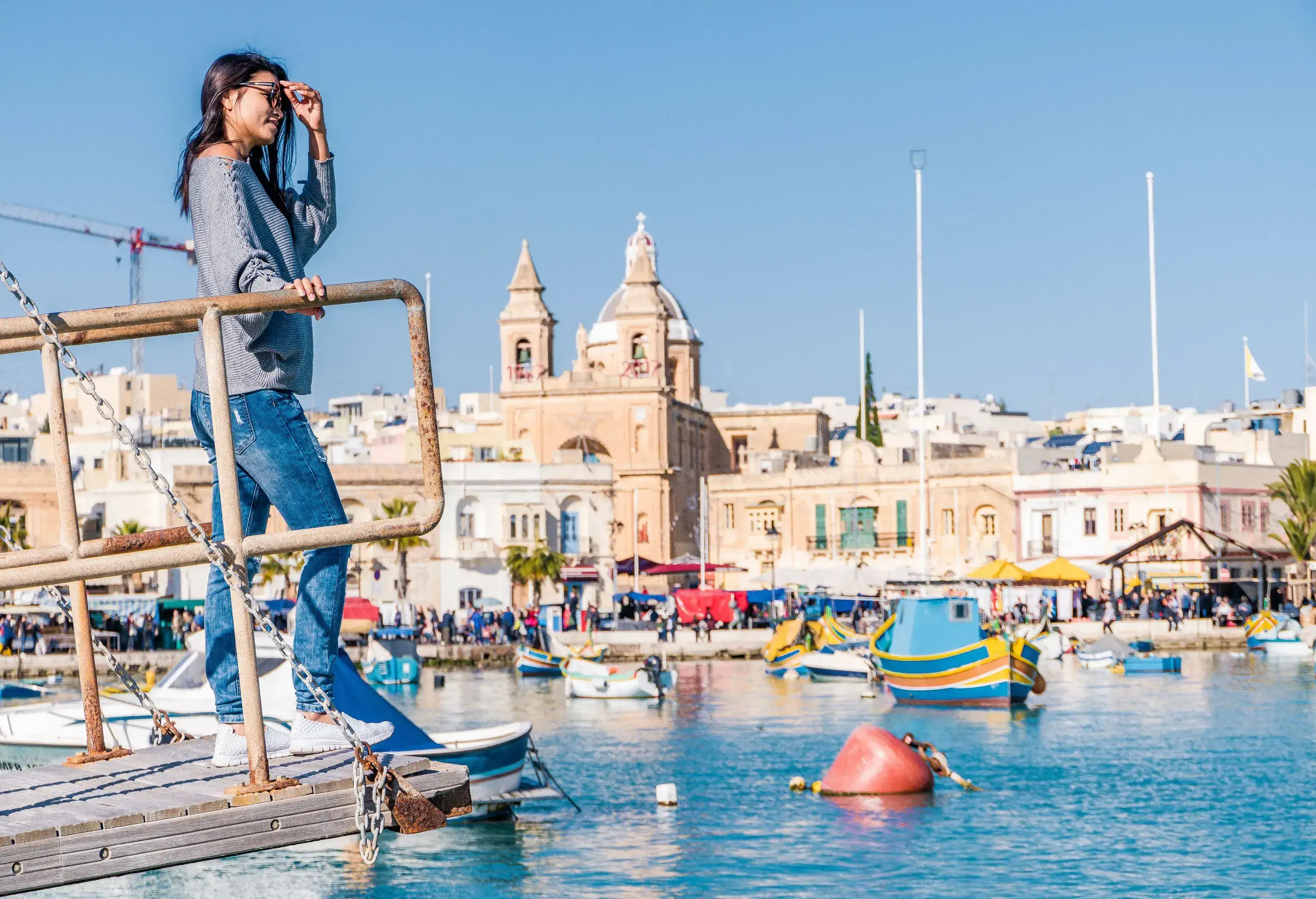 A woman in casual clothes stands on the edge of a bridge overlooking a harbour with anchored boats along a crowded promenade.