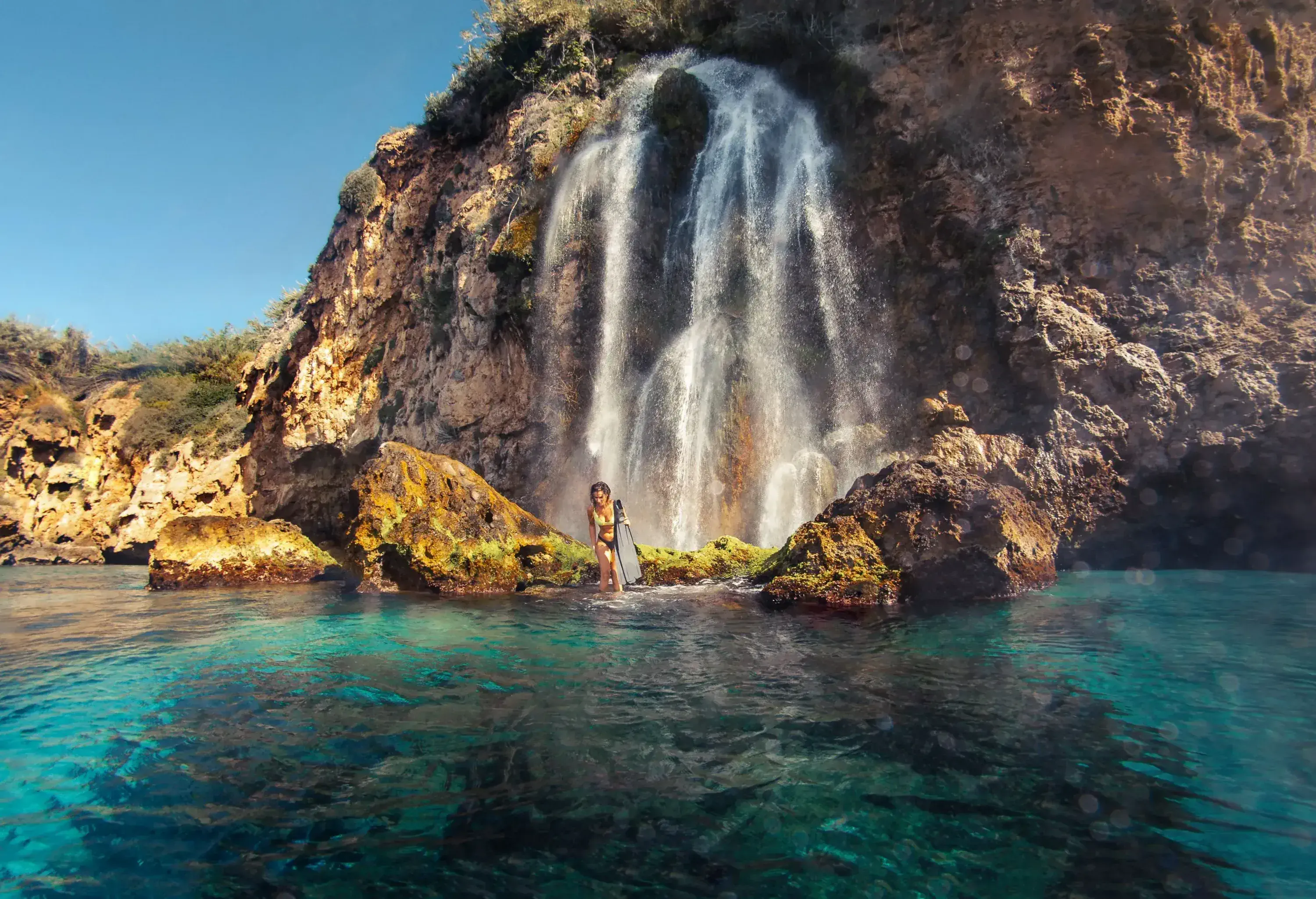 A woman in a bikini walks in the shallow waters under a waterfall while holding a pair of pink flippers.