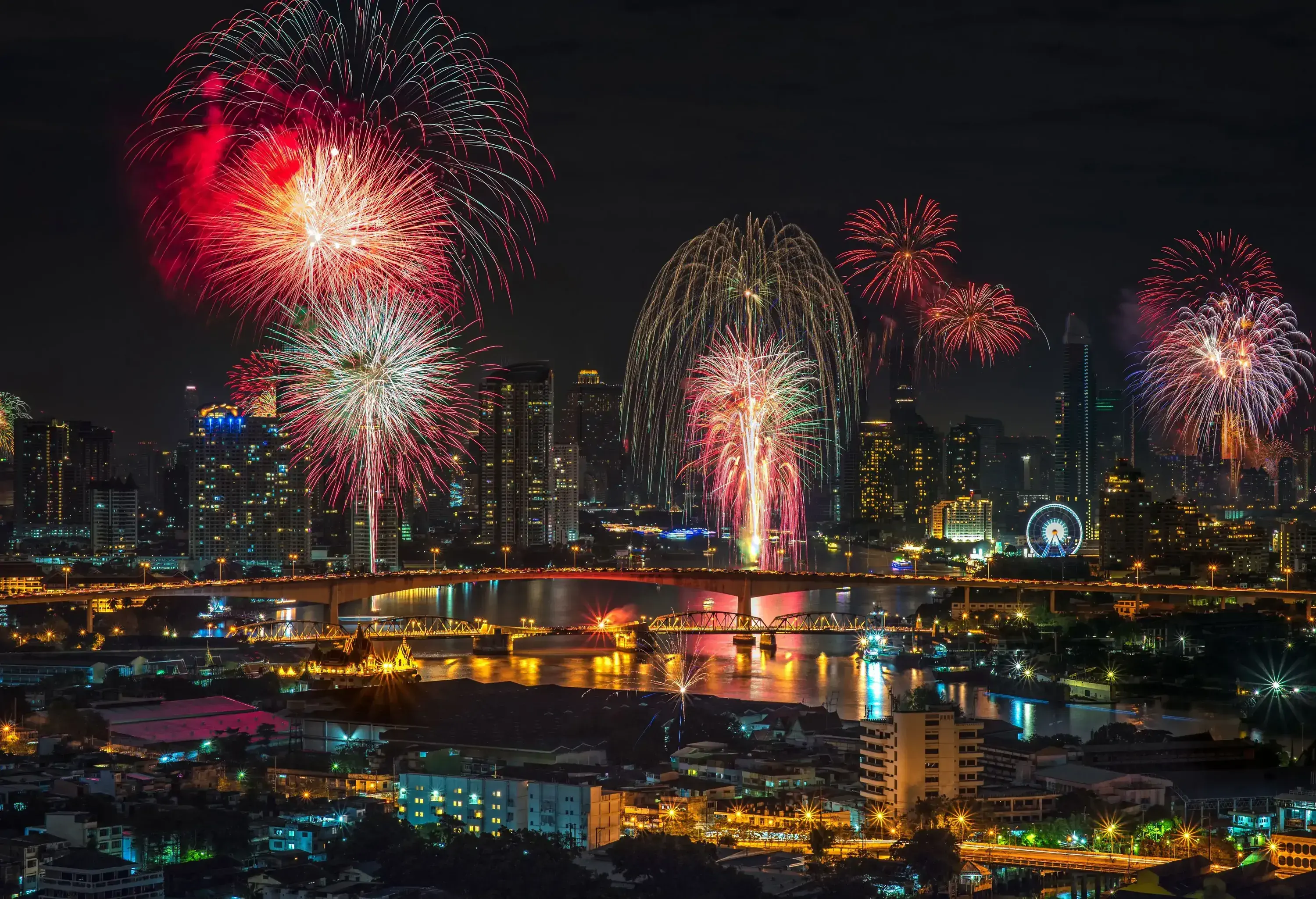 Colourful fireworks above brightly lit urban scenery with skyscrapers and a long bridge above the river.