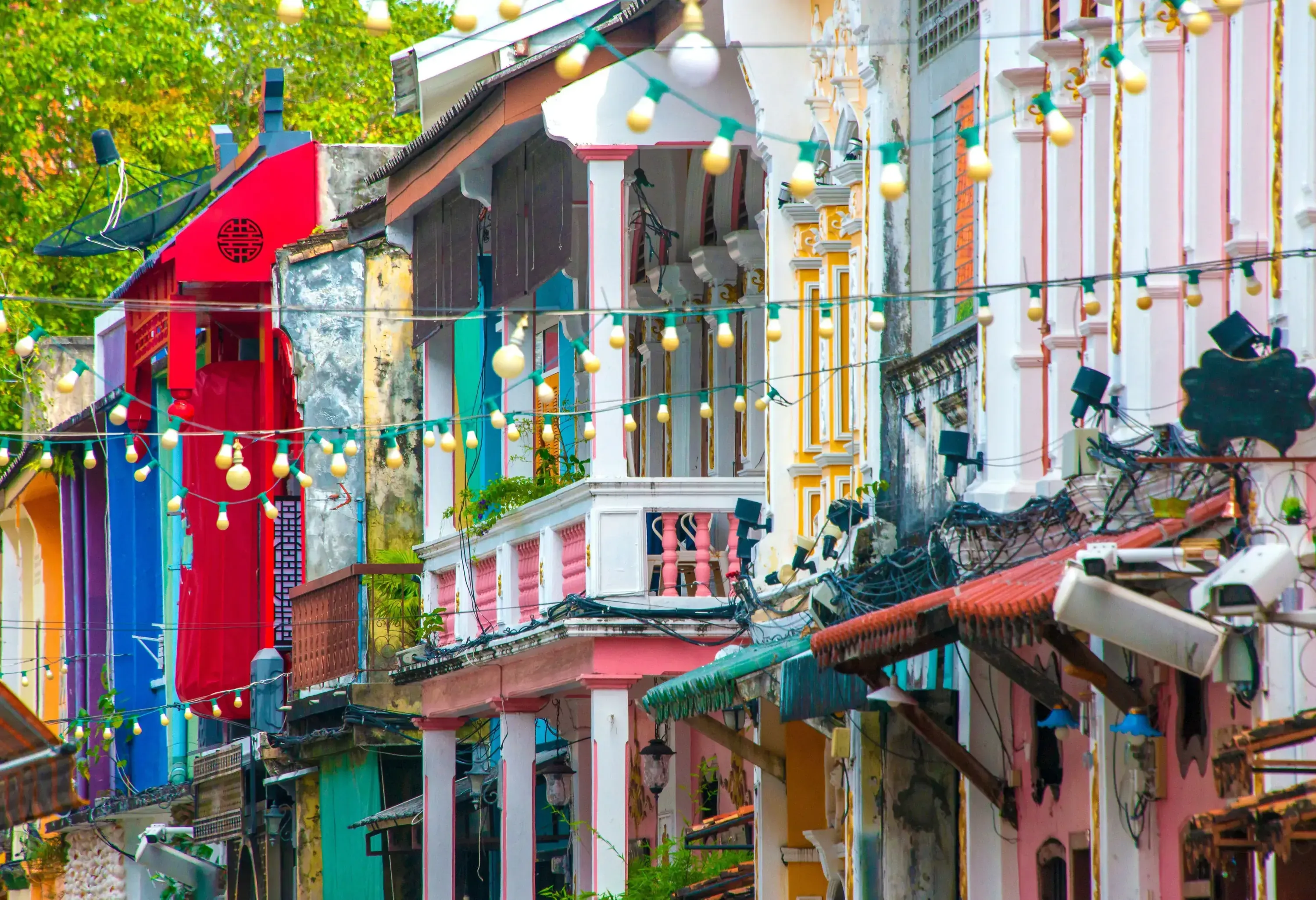 Vibrant, colorful street scene with multicolored historic buildings, ornate details, and strings of warm glowing lanterns. Lush greenery in the background.