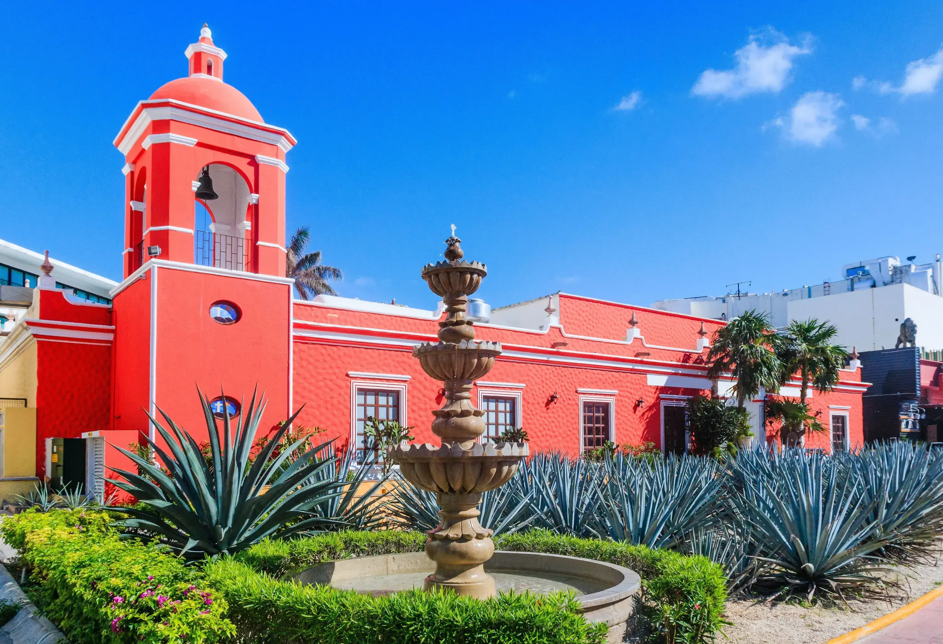 A fountain in a curated garden of aloe vera plants against the backdrop of a pink building with a bell tower.