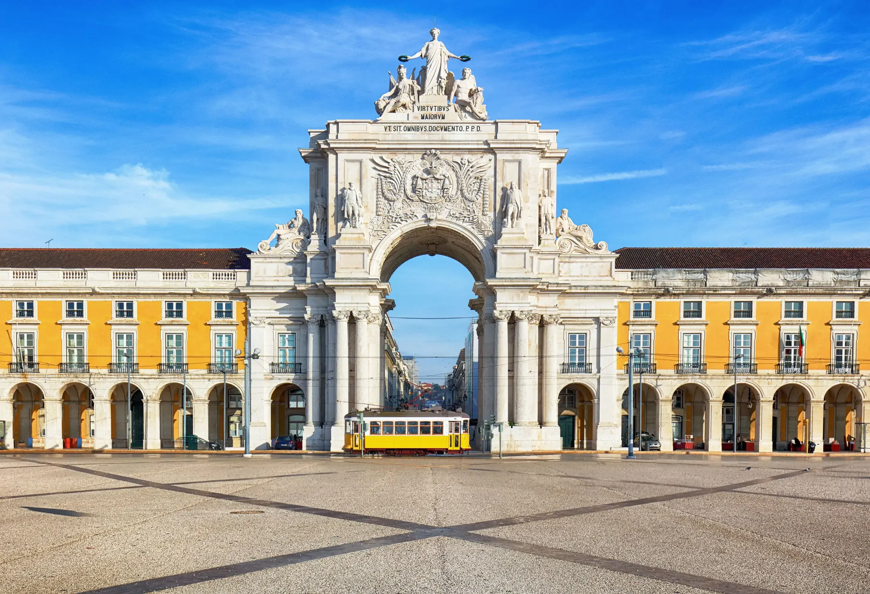 A yellow tram passed the stone memorial arch connecting the arcades supported by columns at regular intervals in a town square.