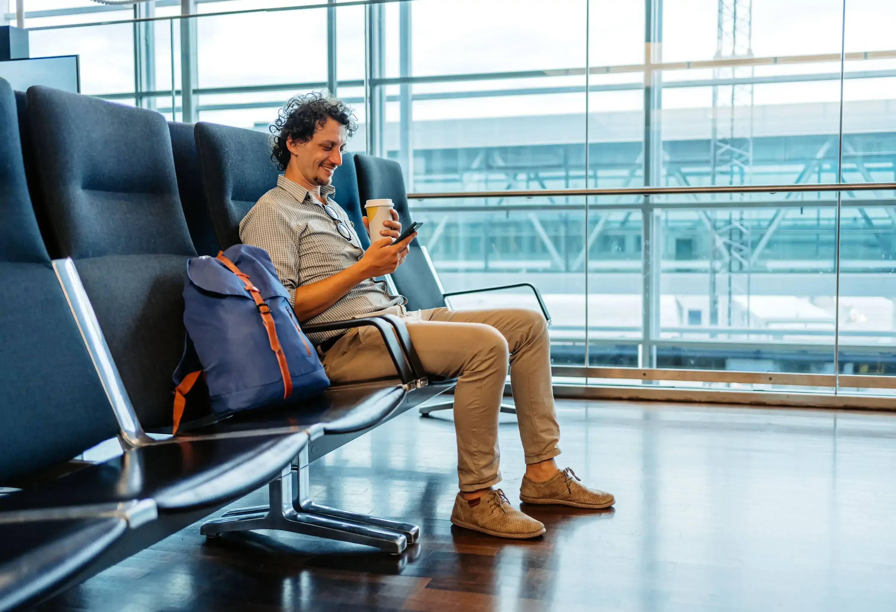 A person sits in a bright airport waiting area while looking at phone and holding a coffee cup, with a backpack on the adjacent seat.
