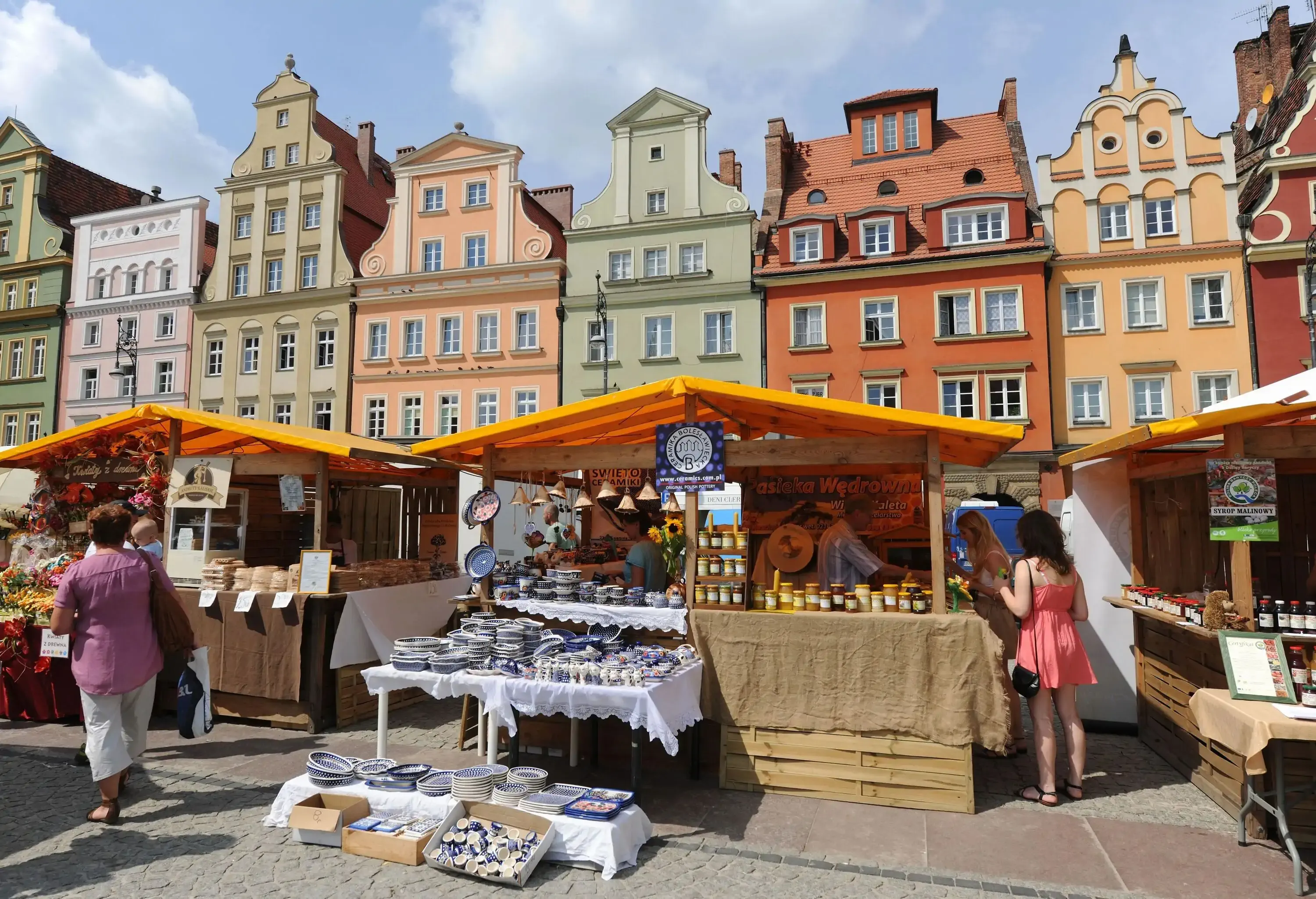 Market stalls with traditional pottery, honey and flowers in a city square against the traditional and colourful buildings in the background.